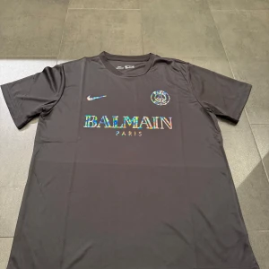 Svart t-shirt från Nike x Balmain Paris - Svart t-shirt från Nike i samarbete med Balmain Paris och PSG. Framsidan har regnbågsskimrande loggor: Nike Swoosh, PSG-emblem och stor Balmain Paris-text. Klassisk passform och rund halsringning.