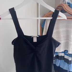 Svart croppad linnetopp - Snygg svart croppad linnetopp med breda axelband och twistad detalj framtill. Perfekt till sommaren och enkel att matcha med olika outfits.