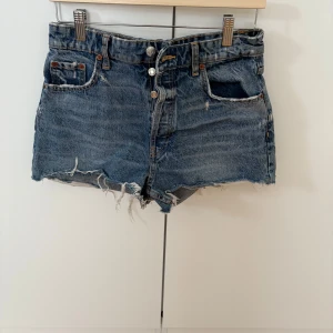 Blå jeansshorts med fransar - Säljer ett par klassiska blå jeansshorts med råa franskantade benslut och låg  midja. Shortsen har fem fickor, knappgylf och en avslappnad passform. Perfekta för sommardagar 