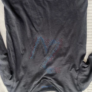 Svart långärmad tröja från Zadig & Voltaire - Cool svart långärmad tröja från Zadig & Voltaire med ett stort glittrigt Z i blått, rött och silver på bröstet. Tröjan har två knappar vid halsen och är i mjuk bomull. Perfekt för dig som gillar statement-plagg!