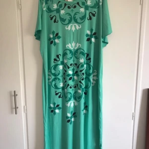 Grön koftan med blommigt mönster - Säljer en lång, lös grön koftan med blommigt mönster i vitt och svart. Koftanen har korta ärmar och ett avslappnat fall, perfekt för varma dagar.