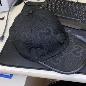 Svart keps från Gucci - Säljer en svart keps från Gucci med diskret GG-mönster över hela kepsen. Kepsen har böjd skärm och är tillverkad i ett slitstarkt tyg. Perfekt accessoar för en stilren look.