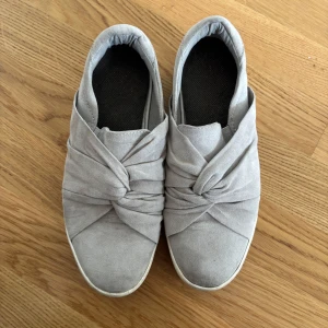Grå slip-on sneakers med knutdetalj - Säljer ett par grå slip-on sneakers med snygg knutdetalj framtill. Skorna har rund tå och platt sula, vilket gör dem bekväma och lätta att ta på. Perfekta för dig som gillar stilrena och enkla skor med en twist. Lite smutsiga därav priset, går såklart att tvätta!