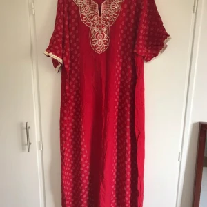 Röd koftan med broderad detalj - Säljer en röd koftan med vita broderade detaljer vid halsringningen och små mönster över hela klänningen. Klänningen har korta ärmar med dekorativ kant och är perfekt för dig som gillar unika plagg med etnisk touch.