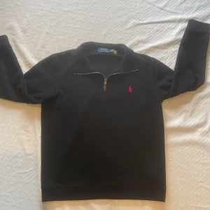 Svart tröja med halv dragkedja från Polo Ralph Lauren - Svart långärmad tröja från Polo Ralph Lauren med halv dragkedja och röd broderad logga på bröstet. Klassisk och stilren design, perfekt för lager-på-lager. Mjuk och bekväm bomullskvalitet.