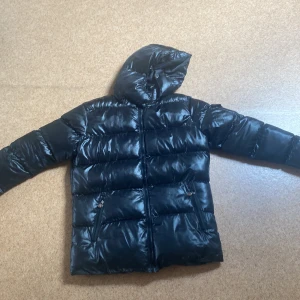 Svart dunjacka från Moncler - Säljer en svart dunjacka från Moncler med huva och dragkedja. Jackan har två snedställda fickor med dragkedja och klassisk Moncler-logga på ärmen. Perfekt för kyliga dagar och riktigt snygg till streetwear.