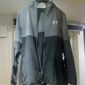Grå och svart vindjacka från Under Armour - Snygg tvåfärgad vindjacka från Under Armour med huva och dragkedja. Jackan är grå upptill och svart nedtill, har lång ärm och reflekterande detaljer. Perfekt för träning eller vardagsstil. Kan gärna byta mot nått också