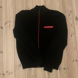 Svart ziptröja från Prada med röd dragkedja - Säljer en svart långärmad ziptröja från Prada med röd dragkedja och logga på bröstet. Tröjan är i bomull och har en stilren design med hög krage. Perfekt för dig som gillar exklusiva märken och snygga detaljer.