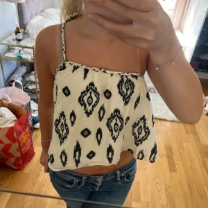 Vit och svart mönstrad croppad linnetopp - Säljer en croppad linnetopp i vitt med svart grafiskt mönster. Toppen har smala axelband och en lös passform, perfekt för varma dagar. Snygg till jeans eller shorts!
