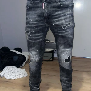 Svarta Dsquared2 jeans med slitningar - Säljer ett par svarta jeans från Dsquared2 med coola slitningar och färgstänk. Jeansen har normal passform, klassisk femficksdesign och knappgylf. Snyggt tvättade detaljer och Dsquared2-logga vid gylfen. Inga problem eller skador alls☺️