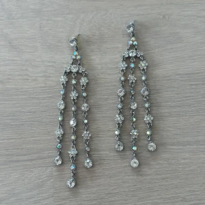 Långa glittriga örhängen med strass - Säljer ett par långa, hängande örhängen i silverfärg med tre rader av gnistrande strass-stenar. Örhängena har ett elegant och festligt utseende med mycket glans och reflektioner. Perfekta för att sticka ut på speciella tillfällen.