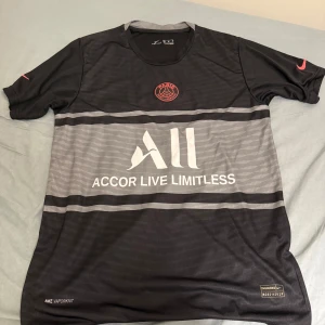 PSG fotbollströja  - Säljer en svart och grå PSG fotbollströja från Nike med Messi #30 på ryggen. Tröjan har korta ärmar, Dri-Fit material och Accor Live Limitless sponsortryck på framsidan. Perfekt för fotbollsfans!