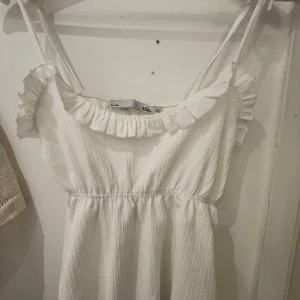 Vit somrigt linne med volang - Söt vitt linne med smala axelband som knyts och volangdetalj vid urringningen. Linnet har en lätt rynkad midja och är tillverkad i ett mjukt, strukturerat tyg. Perfekt för varma dagar och en avslappnad look.