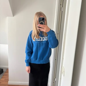 Blå sweatshirt med Brooklyn-tryck - Säljer en blå sweatshirt med vit text där det står 'Brooklyn New York City' på bröstet. Tröjan har en klassisk sweatshirt-passform med muddar vid ärmslut och nederkant. Perfekt för en avslappnad stil.