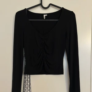 Svart långärmad topp med rynk framtill från Nelly Trend - Svart långärmad topp med djup v-ringning och snygg rynkdetalj framtill från Nelly Trend i storlek XS. 