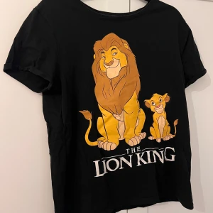 Svart The Lion King t-shirt - Svart t-shirt med stort tryck av Lejonkungen och Simba på framsidan samt texten 'The Lion King' i vitt. Rund halsringning och korta ärmar. Perfekt för dig som älskar Disneyklassiker!