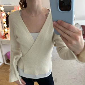 Krämvit omlottkofta från STOCKH LM - Superfin krämvit omlottkofta i mjukt stickat material. Koftan har knyt i midjan och dekorativa guldknappar vid ärmsluten. Perfekt till lager-på-lager och ger en stilren look med sin v-ringning och långa ärmar.