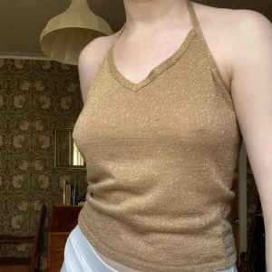 Guldig glittrig halterneck linne - Säljer ett guldig glittrigt halterneck linne med smala axelband som knyts i nacken. Toppen har en croppad passform och är perfekt för dig som vill sticka ut. Materialet är stretchigt och skimrar snyggt i ljuset.