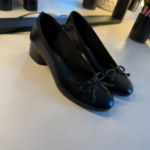 Svarta ballerinaskor med klack - Säljer ett par ballerinaskor med låg klack. Skorna har en söt rosettdetalj framtill. De är använda två gånger. De har även en lite mjukare sula så dem är bekväma. 