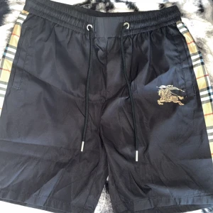 Svarta shorts/badbyxor från Burberry med logga - Svarta Burberry shorts med klassiska rutiga sidopaneler. Perfekta för sommaren med en stilren och exklusiv look. (Material är som badbyxor med funkat också som vanliga shorts och lite mindre passform än M)