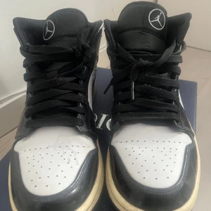 Nike Air Jordan 1 svartvita sneakers - Säljer ett par klassiska Nike Air Jordan 1 sneakers i svart och vitt. Skorna har svart snörning, rund tå och platt sula. Ikonisk Air Jordan-logga på sidan och Nike swoosh. Perfekta för dig som gillar streetwear och vill ha en tidlös look.