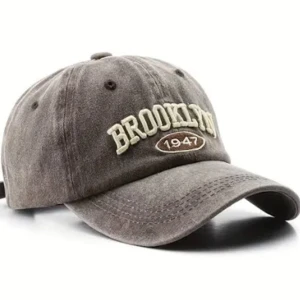 Grå keps med BROOKLYN-brodyr - Snygg grå keps med böjd skärm och broderad text 'BROOKLYN 1947' framtill. Justerbar rem baktill för perfekt passform. Perfekt accessoar för en avslappnad stil.