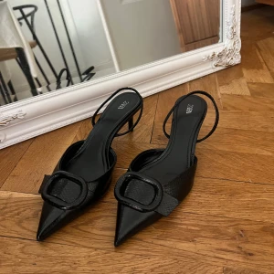 Svarta klackskor med spetsig tå från Zara - Enbart använda en gång! Säljer ett par eleganta svarta klackskor från Zara med spetsig tå och stor dekorativ spänne framtill. Skorna har en tunn rem runt hälen och är tillverkade i glansigt material med struktur. Perfekta för att lyfta din outfit.