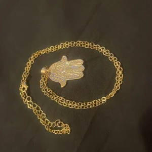 Guldigt halsband med Hamsa-hänge (fatimas hand) - Säljer ett snyggt halsband i guld med ett detaljerat Hamsa-hänge dekorerat med små glittrande stenar. Kedjan är tunn och elegant, perfekt för att ge din outfit en unik touch.