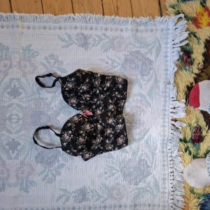 Svart blommig korsett-topp med rosett - Supersöt svart korsett-topp med små blommor i rosa och grönt samt en rosa rosett framtill. Justerbara axelband och lätt vadderade kupor. Perfekt för att lyfta din outfit med en romantisk touch.