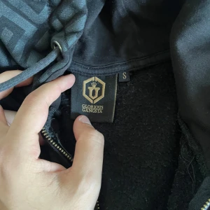 Svart hoodie från Glorious Gangsta med mönster - ÄKTA✅Säljer en svart hoodie från Glorious Gangsta med dragkedja och huva. Jackan har ett grått geometriskt mönster upptill och guldfärgade detaljer på dragkedjor och tryck. Tillverkad i polyester och bomull.