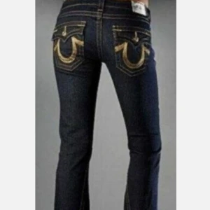 W26 True Religion mörkblå bootcut jeans med guldiga detaljer - Ovanliga vintage mörkblå jeans från True Religion med bootcut-modell. Byxorna har låg midja och ikoniska guldiga broderade detaljer på bakfickorna. Klassisk femficksdesign och snygga kontrastsömmar. Perfekta för dig som gillar statement-jeans! Storlek W26. Innerbenslängd: 71 cm. Ytterben: 91 cm. 