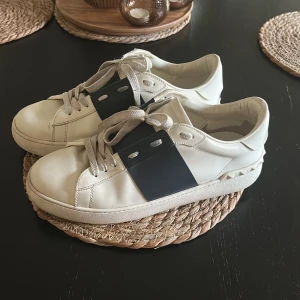 Valentino opens skor - Säljer ett par vita Valentino sneakers med en bred svart rem över sidan. Skorna har snörning och platt sula, samt diskreta nitar baktill. Perfekta för dig som gillar stilrena och exklusiva sneakers.