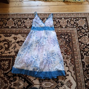 Blommig sommarklänning med blå detaljer - Söt och luftig klänning med blommigt mönster i blått och lila på vit botten. Klänningen har v-ringning, breda axelband och markerad midja med blått band samt blå kant nertill. Perfekt för varma dagar.