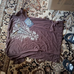 Brun t-shirt med tryck - Säljer en brun t-shirt med detaljerat tryck i vitt, gult och rött framtill. T-shirten har korta ärmar och rund hals. Perfekt för dig som vill ha en unik och snygg stil.