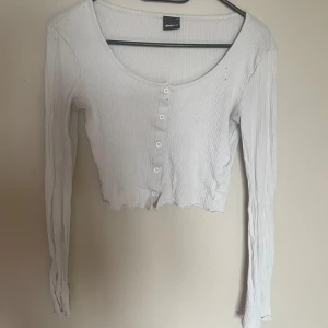 Vit ribbad croppad topp från Gina Tricot - Säljer en vit ribbad croppad topp från Gina Tricot med långa ärmar och knappar framtill. Toppen har en rundad urringning och är tillverkad i ett stretchigt material som sitter snyggt på kroppen.🤍