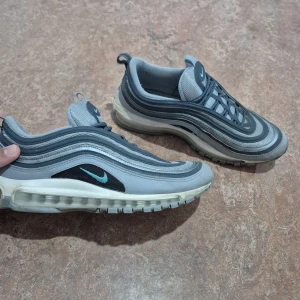 Nike Air Max 97  - Nike Air Max 97 sneakers i grå och ljusblå färg med turkos Swoosh. Skon har den klassiska vågiga designen, synlig luftdämpning i sulan och snörning. Perfekt för dig som gillar streetwear och sportig stil.