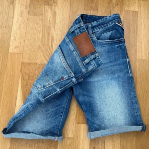 Blå jeansshorts från Replay - Snygga blå jeansshorts från Replay med klassisk femficksdesign och ljusa slitningar. Shortsen har normal passform, knappgylf och läderpatch med logga bak i midjan. Perfekta för varma dagar när du vill ha en avslappnad stil.