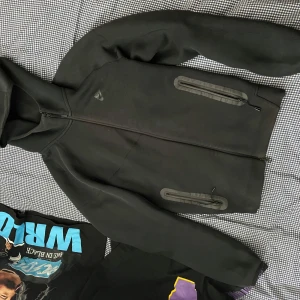 Svart softshelljacka med huva från Nike - Säljer en svart softshelljacka från Nike med huva och hel dragkedja. Jackan har två vattentäta fickor med dragkedja framtill och en sportig look. Perfekt för dig som gillar stilren och funktionell design.