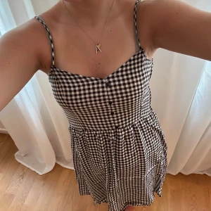 Svartvit rutig klänning med smala axelband - Superfin kort klänning med svartvitt rutigt mönster och smala axelband. Klänningen har knappar framtill och en lätt utställd kjol. Perfekt för varma dagar och enkel att styla med sneakers eller sandaler.