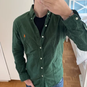 Grön skjorta från Ralph Lauren - Säljer en snygg grön skjorta från Ralph Lauren i slim fit-modell. Skjortan har klassisk krage, knappar framtill och den ikoniska broderade loggan i orange på bröstet. Perfekt till jeans eller chinos.