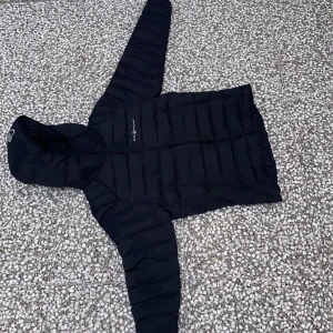 Svart dunjacka med huva från Allied Feather + Down - Säljer en svart dunjacka med huva från Allied Feather + Down. Jackan har quiltad design och är lätt men varm tack vare 800 fill power dun. Perfekt för kyliga dagar och enkel att bära med sig.