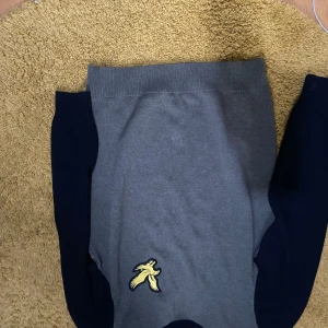 Grön tröja med mörkblå ärmar från Lyle & Scott - Snygg grå tröja med mörkblå ärmar från Lyle & Scott. Tröjan har en gul broderad logga framtill och ribbade muddar vid hals och ärmslut. Perfekt för en stilren och avslappnad look.