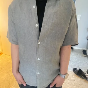 Grå linneskjorta med kort ärm - Snygg olivgrön skjorta i linne med korta ärmar och vita knappar. Skjortan har en avslappnad passform och är perfekt för varma dagar. Enkel och stilren design som passar till många olika outfits. Köptes för 400kr, använd en gång!