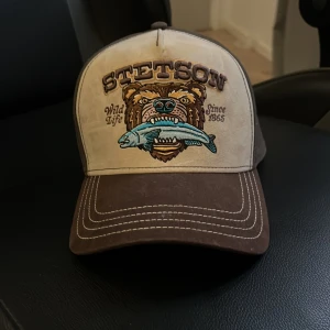 Beige och brun keps från Stetson - Säljer en beige och brun keps från Stetson med broderad björn och fisk på framsidan samt texten 'Wild Life Since 1865'. Justerbar baktill med plastspänne. Perfekt för dig som gillar naturinspirerad stil. Helt oanvänd 