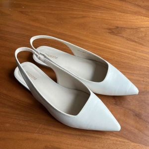 Vita ballerinaskor från Graceland - Säljer ett par stilrena vita ballerinaskor från Graceland med öppen häl och spetsig tå. Skorna har en elegant design och är super fina! Liten slitning framme på tån men knappt använda!🥰Passar perfekt till studenten eller annat🙈