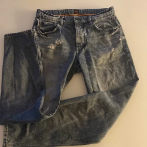 Blå jeans från BOSS med slitningar - Säljer ett par blå jeans från BOSS med snygga slitningar framtill. Klassisk femficksmodell med normal passform och raka ben. Jeansen har knapp- och dragkedjegylf samt tydliga detaljer vid fickorna.