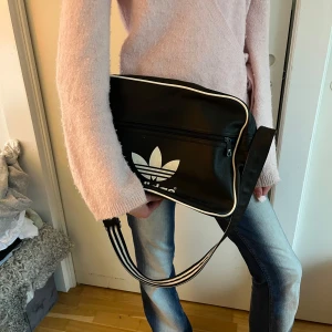 Svart axelväska från Adidas💕🌺 - Säljer en svart axelväska från Adidas med vit logga och vita detaljer. Väskan har ett stort fack och en dragkedjeficka framtill. Perfekt för att bära med sig det viktigaste till skolan eller stan.🌺
