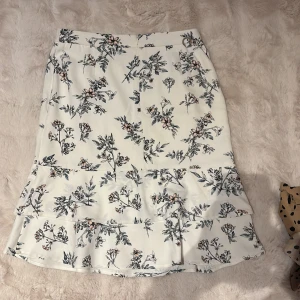 Vit blommig kjol från Stockholm  - Superfin vit kjol med blommigt mönster i svart och orange från Stockholm . Kjolen har en lätt utsvängd form och volang nertill, perfekt för en fräsch och somrig look.