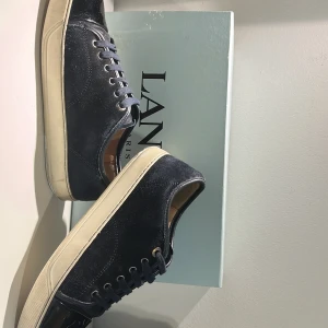 Svarta sneakers från Lanvin i mocka - Säljer ett par svarta Lanvin sneakers i mocka med vita sulor och svarta snören. Skorna har rund tå och klassisk design med diskret logga på sulan. Perfekta för dig som gillar stilrena och lyxiga sneakers.