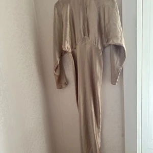 Beige långklänning med lång ärm - Säljer en beige långklänning med lång ärm och dragkedja baktill. Klänningen har en lös passform upptill och smalnar av nedtill. Tillverkad i ett mjukt, syntetiskt material. Perfekt för dig som gillar minimalistisk stil.